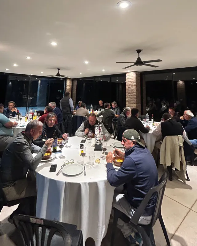 Guests having dinner at round tables with white tablecloths in a glass-walled hall, at night