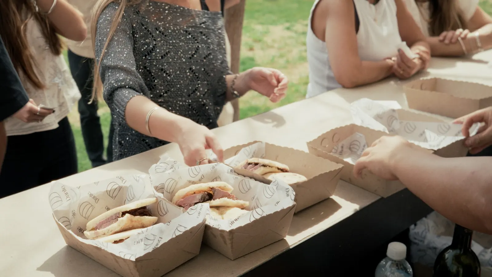 Standing guests take picanha sandwiches with dips, served in paper boats on a garden table