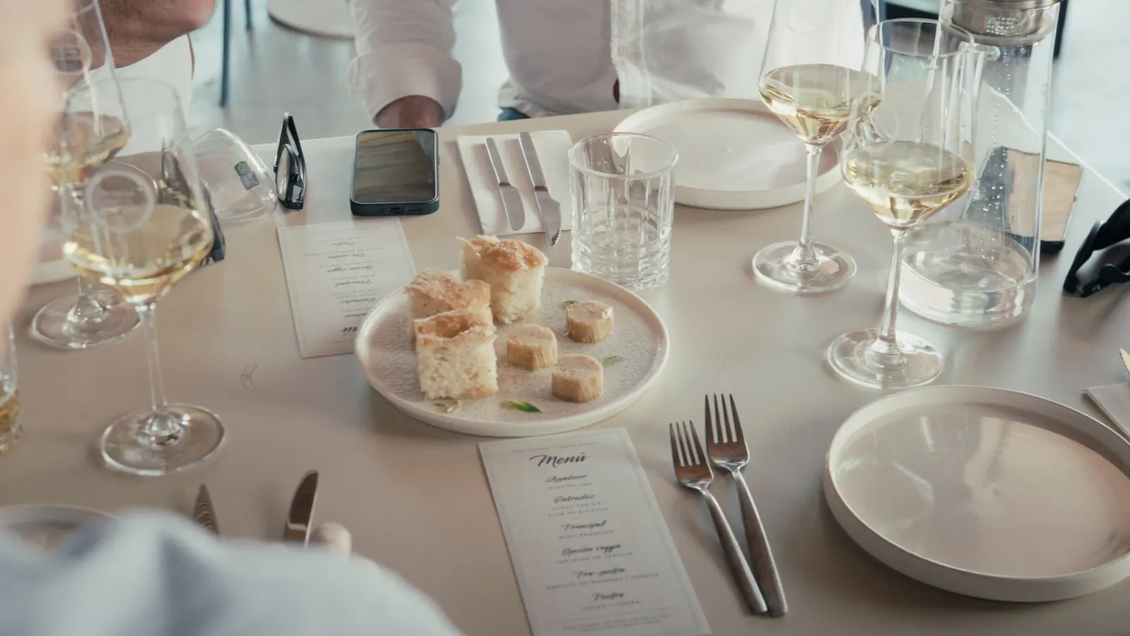 Plate of focaccia squares and rounds of brown butter on the set table, next to the printed menu and glasses of white wine
