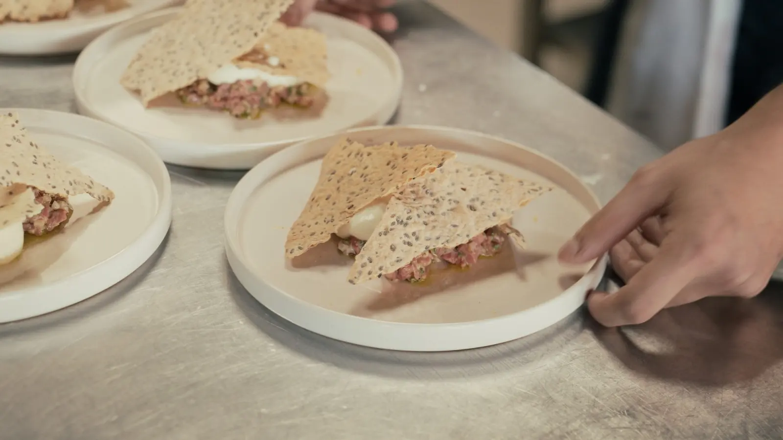 Plates of beef tenderloin tartare under crisp seed tuiles on a steel counter